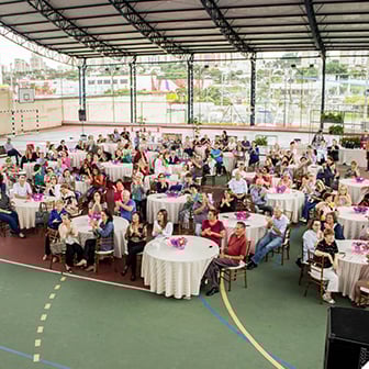 Imagem de uma festa em um ginásio, com muitas crianças e adultos sentados, em um ambiente com iluminação natural.