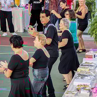 Um grupo de adultos e idosos dança em um evento escolar ao ar livre, com um homem segurando um microfone.