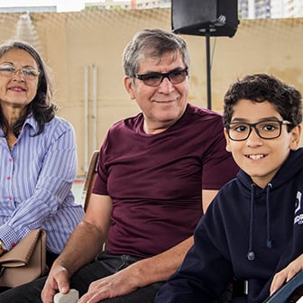 Homem de óculos escuros sorrindo ao lado de mulher de camisa listrada e menino de moletom azul com óculos, sentados.