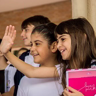 Duas alunas sorridentes com uniformes brancos em um corredor. Uma delas acena e segura um caderno rosa.