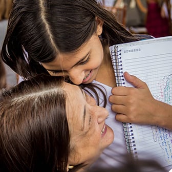 Uma menina sorridente abraça com afeto uma mulher mais velha, segurando um caderno escolar.
