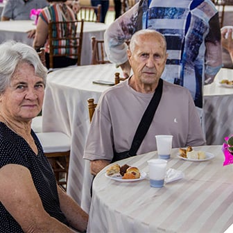 Um casal de idosos sentado em uma mesa durante um evento no Poliedro, com pratos de salgados e doces.