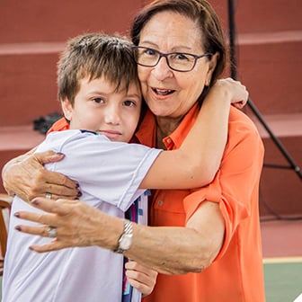 Uma mulher mais velha de óculos e camisa laranja abraça com carinho um jovem estudante uniformizado em um evento escolar.