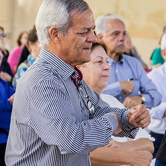 Um homem idoso de camisa listrada participa de uma atividade em grupo ao ar livre, com outros adultos e idosos ao fundo.