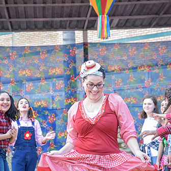 Mulher sorridente vestida com roupa de festa junina, rodeada de crianças em trajes típicos, em decoração de festa junina.