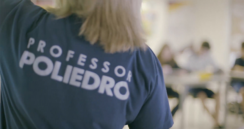 Vista das costas de uma professora com cabelo loiro usando uma camiseta azul com a estampa branca "PROFESSOR POLIEDRO".